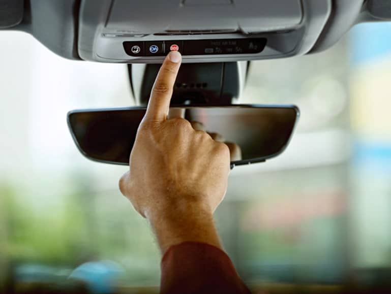 A person is pressing a red button on the overhead console inside a car. The rearview mirror is visible below the hand. The background is blurred, suggesting the car is in a bright, daylight setting.