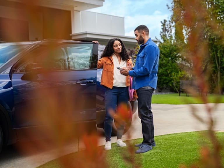 A woman and man stand by a dark blue SUV parked on a driveway. The woman holds the car door open, while the man points at something on a tablet. Blurred branches in the foreground frame the scene.