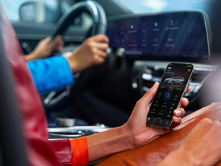 A person in a car is holding a smartphone, displaying a car control app. The person wears a blue shirt and is steering the car with their left hand. The car’s dashboard is visible, showing various controls and displays.