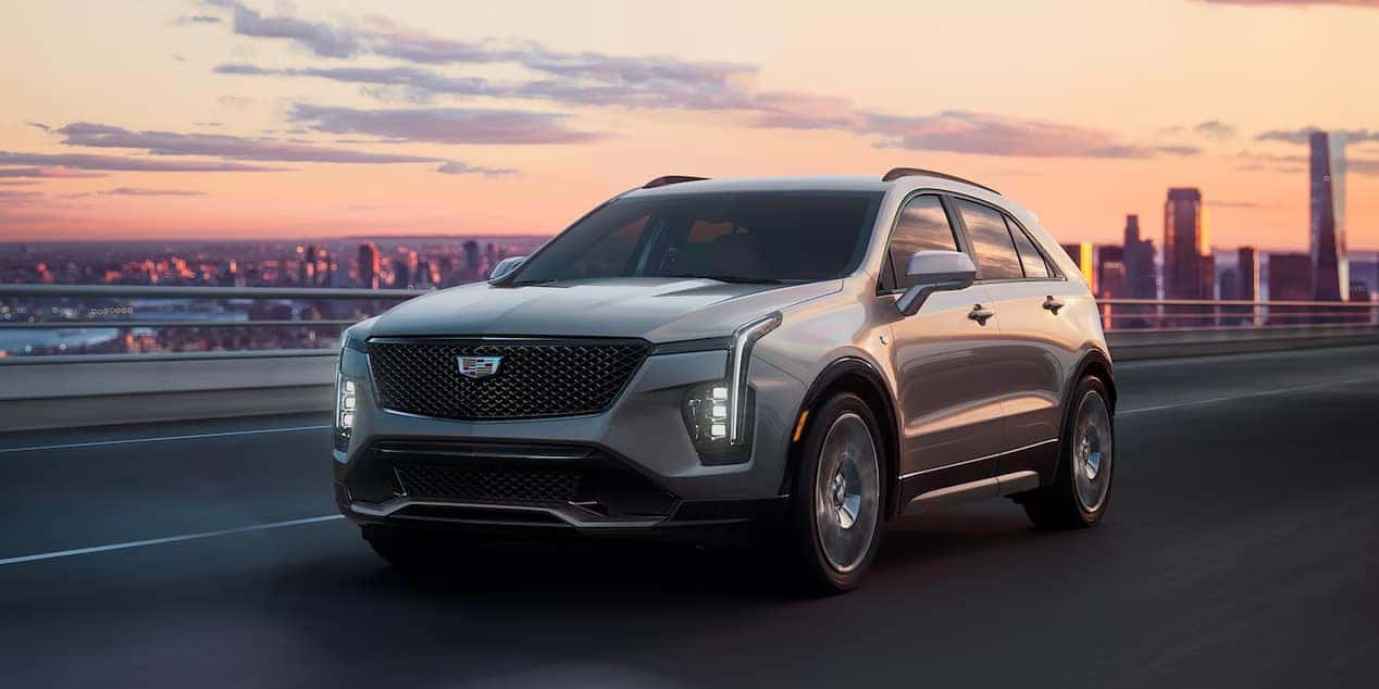 Silver SUV parked on rocky terrain with greenery and mountain range in the background. The SUV is facing slightly to the right, showcasing its front and side profile. The sky is clear and blue.