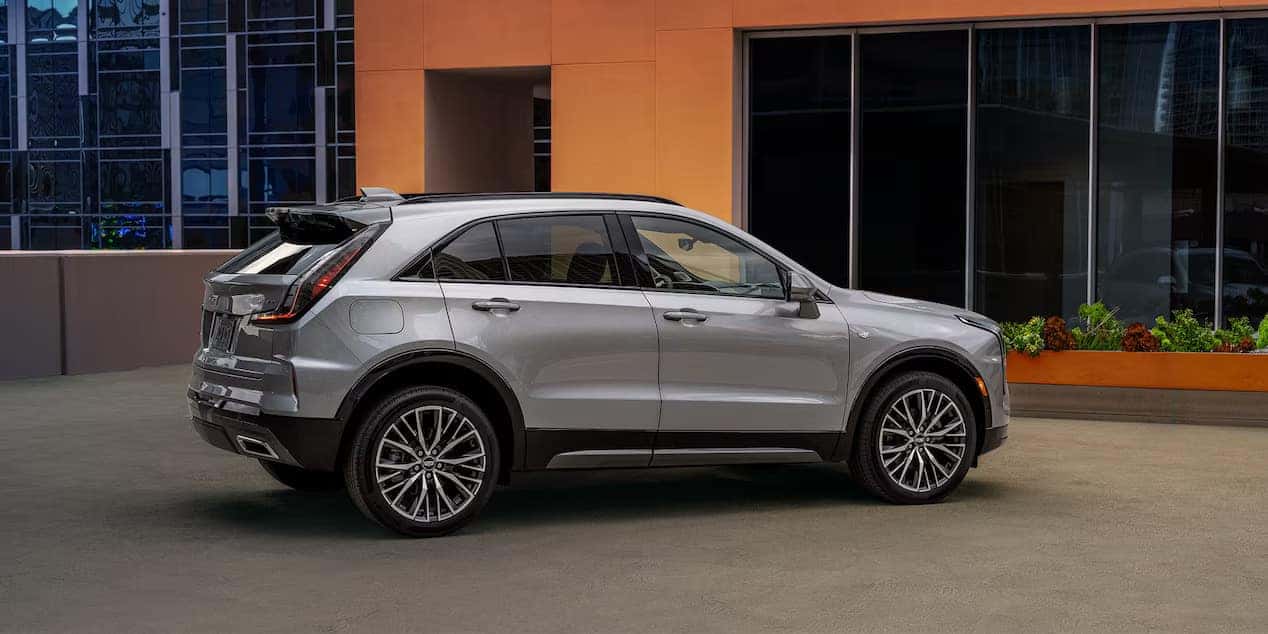 A silver SUV parked on a rocky, mountainous terrain with a scenic view of green hills and a clear blue sky in the background. Yellow flowers are visible among the rocks in the foreground.