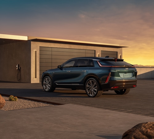 A silver SUV parked on a rocky, mountainous terrain with a scenic view of green hills and a clear blue sky in the background. Yellow flowers are visible among the rocks in the foreground.