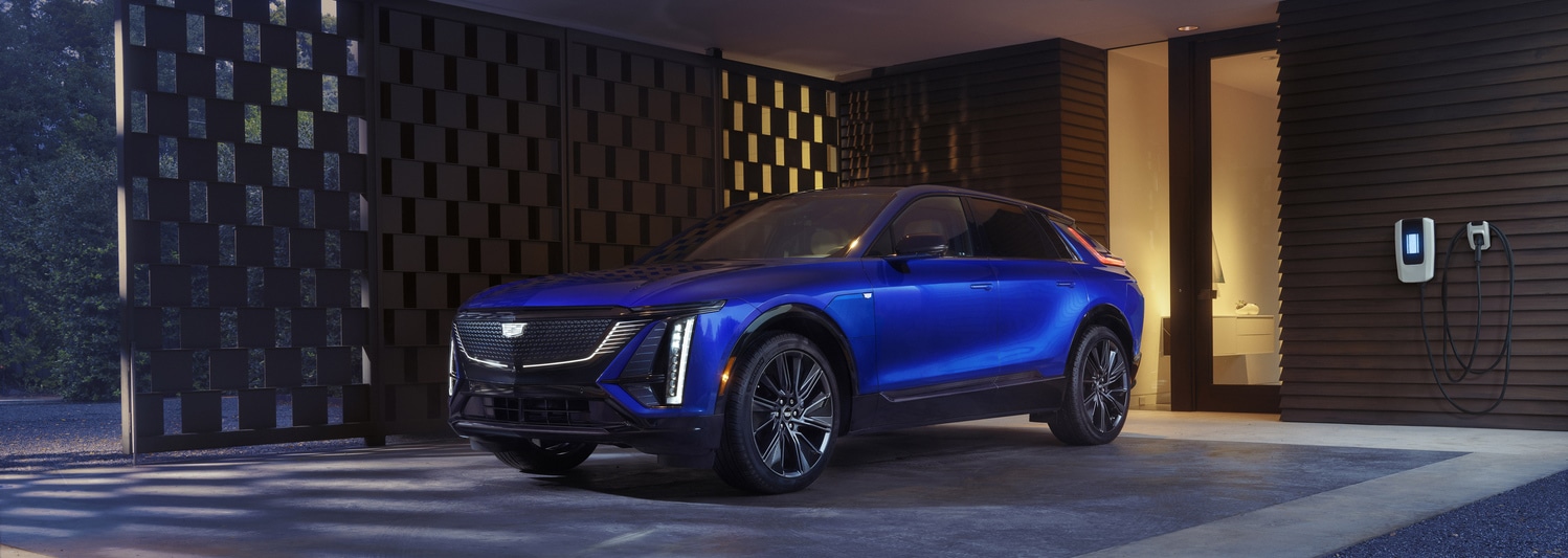 Silver SUV parked on rocky terrain with greenery and mountain range in the background. The SUV is facing slightly to the right, showcasing its front and side profile. The sky is clear and blue.