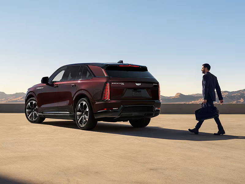 Man walking towards a sleek, maroon crossover SUV parked on a sunny rooftop with a mountainous backdrop.